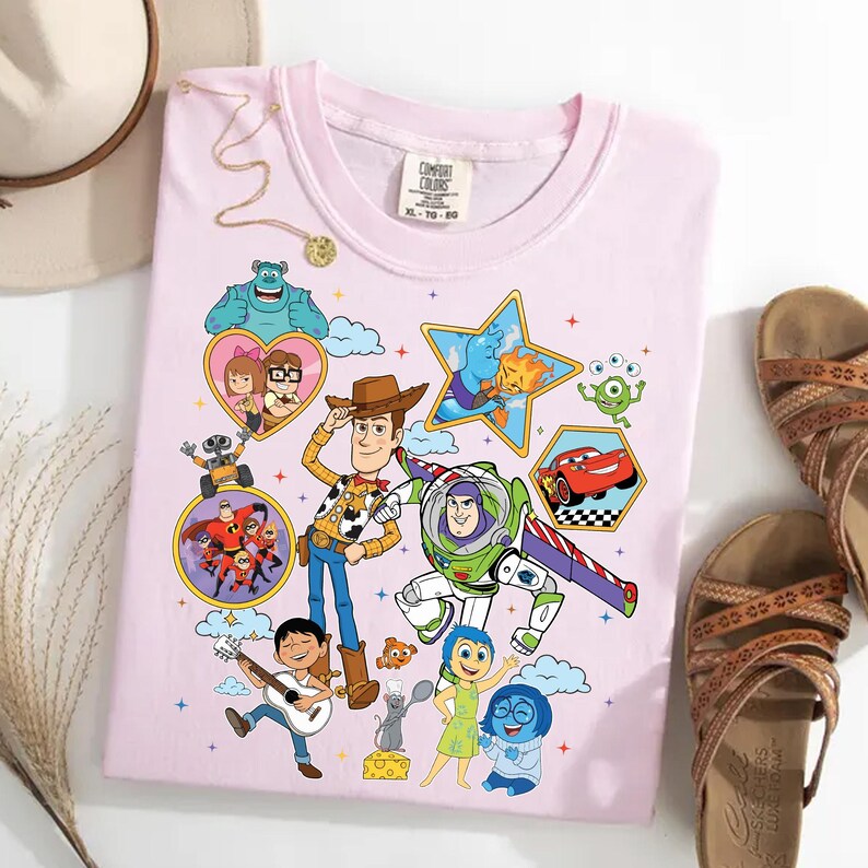 May include: A light pink t-shirt featuring colorful cartoon characters from various animated movies. The design includes characters like Woody, Buzz Lightyear, and others, with the Comfort Colors logo visible.