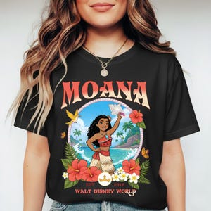 May include: Black t-shirt featuring a colorful graphic of Moana from Walt Disney World. The design includes the word "MOANA" in orange, a circular image of Moana on a beach, and floral accents. "EST 2016" and "WALT DISNEY WORLD" are also printed.