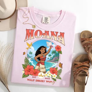 May include: A light pink t-shirt featuring the word "MOANA" in an orange and white font. The shirt has a graphic of Moana holding a seashell, surrounded by flowers and butterflies. The text "EST 2016 WALT DISNEY WORLD" is also printed on the shirt.