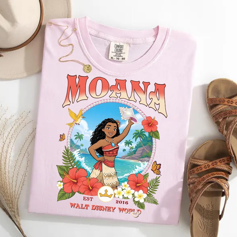 May include: A light pink t-shirt featuring the word "MOANA" in an orange and white font. The shirt has a graphic of Moana holding a seashell, surrounded by flowers and butterflies. The text "EST 2016 WALT DISNEY WORLD" is also printed on the shirt.