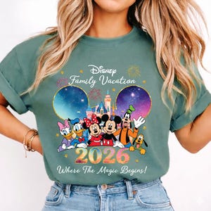 May include: Sage green t-shirt featuring Disney characters Mickey, Minnie, Donald, Daisy, Goofy, and Pluto. The shirt has the text "Disney Family Vacation 2026 Where The Magic Begins!" with a castle and fireworks design.