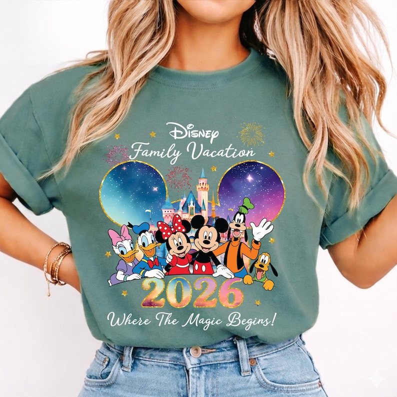 May include: Sage green t-shirt featuring Disney characters Mickey, Minnie, Donald, Daisy, Goofy, and Pluto. The shirt has the text "Disney Family Vacation 2026 Where The Magic Begins!" with a castle and fireworks design.