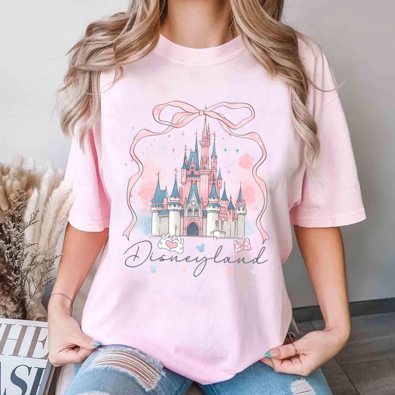 Op de afbeelding: Lichtroze T-shirt met een pastelkleurige illustratie van een sprookjeskasteel met roze en blauwe torens, een roze lint en het woord "Disneyland" in script. Het shirt is gemaakt van een zacht, comfortabel materiaal.