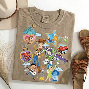 May include: A tan Comfort Colors t-shirt featuring colorful cartoon characters from various animated movies. The design includes Woody, Buzz Lightyear, and other characters in a playful arrangement. The shirt is paired with sandals and accessories.