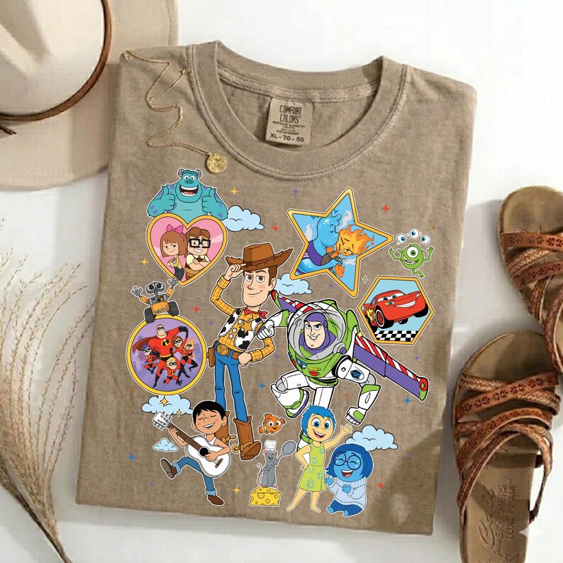 May include: A tan Comfort Colors t-shirt featuring colorful cartoon characters from various animated movies. The design includes Woody, Buzz Lightyear, and other characters in a playful arrangement. The shirt is paired with sandals and accessories.