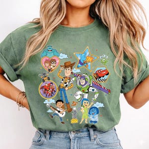 May include: Green t-shirt featuring a colorful graphic of various animated characters. The design includes Woody, Buzz Lightyear, Sulley, and other characters from popular animated movies. The shirt is made of a soft material.