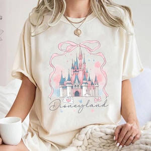 Op de afbeelding: Cr&egrave;mekleurig T-shirt met een Disneyland kasteelontwerp in roze en blauw, met een roze lint en het woord "Disneyland". Het shirt heeft korte mouwen en een losse pasvorm.