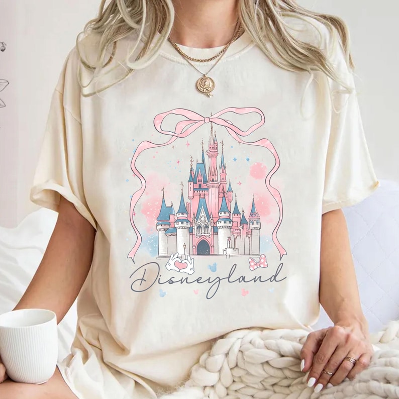 Op de afbeelding: Cr&egrave;mekleurig T-shirt met een Disneyland kasteelontwerp in roze en blauw, met een roze lint en het woord "Disneyland". Het shirt heeft korte mouwen en een losse pasvorm.