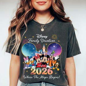May include: Dark gray t-shirt featuring Disney characters Mickey, Minnie, Donald, Goofy, and Pluto. The shirt has the text "Disney Family Vacation 2026 Where The Magic Begins!" with fireworks and castle graphics.