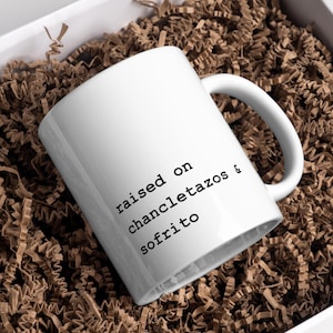 Puede incluir: Una taza de cerámica blanca con el texto "raised on chancletazos & sofrito" en letras negras. La taza está dentro de una caja blanca llena de papel de seda marrón.