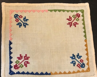 May include: A white linen square with a blue, green, pink, and orange border. The center of the square has four embroidered floral designs in red, green, and blue.