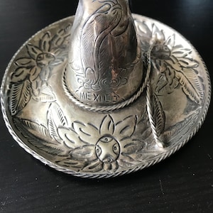 May include: A silver sombrero with intricate floral designs and the word "MEXICO" inscribed on the brim.