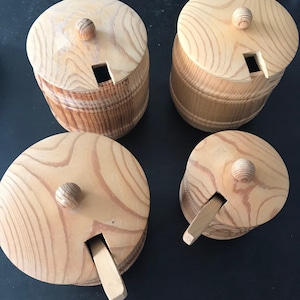 Puede incluir: Cuatro tarros de miel de madera con tapas y una pequeña cuchara de madera. Los tarros son de madera marrón claro con un patrón de veta natural.