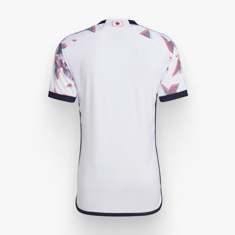 Op de afbeelding: Wit voetbaltenue met een blauw en rood geometrisch patroon op de mouwen. Het tenue heeft een donkerblauwe rand rond de kraag en mouwen. De Japanse vlag bevindt zich op de achterkant van de kraag.