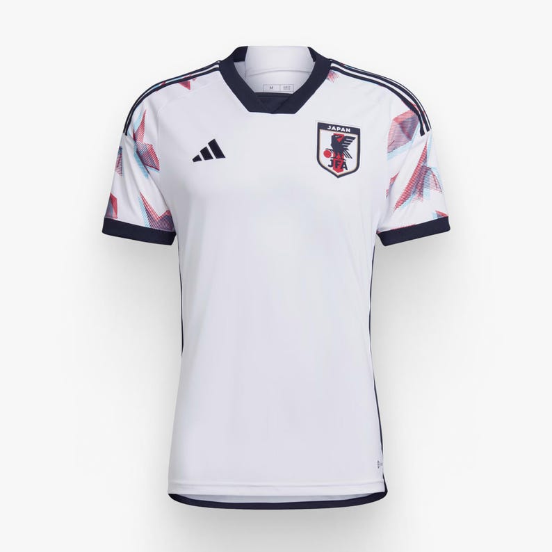Op de afbeelding: Wit voetbaltenue met blauwe en rode accenten. Het tenue heeft een geometrisch patroon en bevat het Adidas-logo en het embleem van het Japanse nationale team met de tekst "JAPAN" en "JFA".