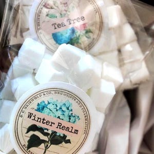 May include: A close-up of white, cube-shaped items, possibly soaps or bath bombs, packaged in clear cellophane. Two circular labels are visible, one reading "Tea Tree" and the other "Winter Realm", with floral and botanical illustrations.