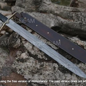 Può includere: Una spada a doppio taglio con lama in acciaio Damasco a motivi, impugnatura rivestita in pelle nera e guardia metallica. La spada è accompagnata da un fodero in pelle marrone scuro con rivetti metallici color argento lungo la lunghezza.