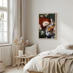 May include: A framed print of a woman with long red hair sleeping with a tiger. The woman is wearing a white dress and the tiger is lying on top of her. The background is a lush green forest with white flowers.