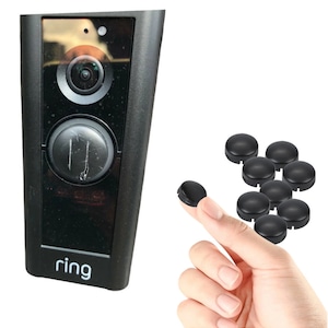 May include: A black Ring doorbell with a round button and a camera lens. The doorbell is mounted on a white wall. There are 10 black plastic covers for the doorbell button.