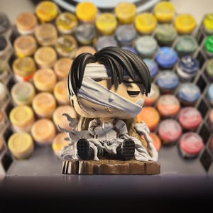 Puede incluir: Una figura de un personaje con cabello negro, una venda blanca sobre los ojos y un atuendo blanco. La figura está sentada sobre una base marrón. El fondo presenta numerosos botes de pintura pequeños.