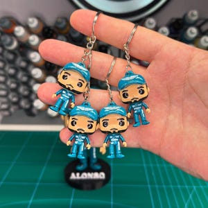 Puede incluir: Cuatro llaveros azules y blancos con una figura de dibujos animados con un casco azul y un traje de carreras blanco. Los llaveros están unidos a llaveros de plata. Los llaveros están sobre una superficie verde con la palabra "ALONSO" impresa en un círculo negro.