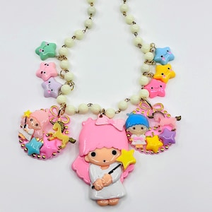 May include: A whimsical necklace featuring a series of colorful star charms and character pendants. The necklace has a cream-colored beaded chain with gold-tone accents. The pendants include a pink character with a star wand.