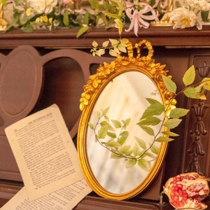 May include: An ornate, gold-framed oval mirror with a crown detail, reflecting a room with flowers and greenery. The mirror rests on a dark wooden surface, with book pages and a halved pomegranate nearby. The mirror's frame is embellished.
