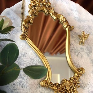 Ornate baroque-style gold mirror with intricate floral carvings and chain, styled on lace fabric with green leaves and a mini cherub sculpture beside it.