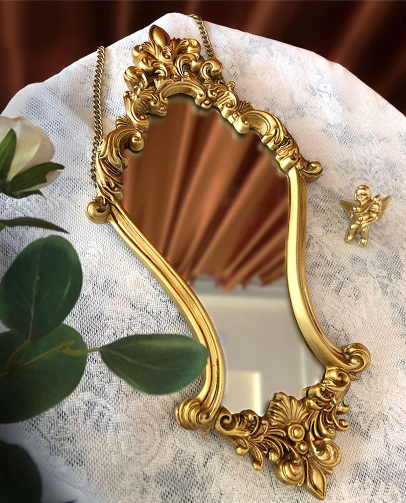 Ornate baroque-style gold mirror with intricate floral carvings and chain, styled on lace fabric with green leaves and a mini cherub sculpture beside it.