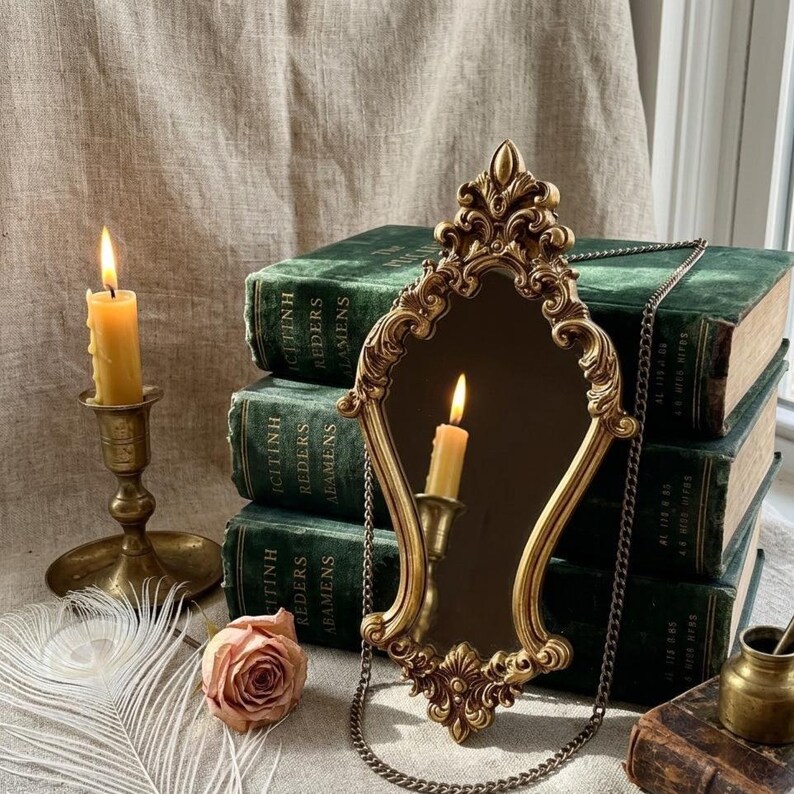 A vertical 20 x 40 cm gold Rococo hourglass wall mirror leaning against dark green velvet antique books on a linen surface. This ornate Baroque vanity decor features detailed golden scrollwork and a single hanging metal chain. A flickering candle is reflected in the glass, surrounded by a blush rose, a white peacock feather, and a vintage brass inkwell. The warm lighting highlights the hand-painted patina and the elegant, slender silhouette.