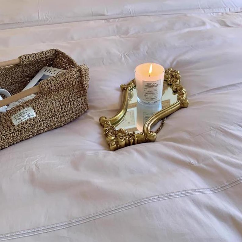 Small gold ornate mirror used as a tray on a bed, holding a lit white candle next to a woven basket filled with books and mugs.