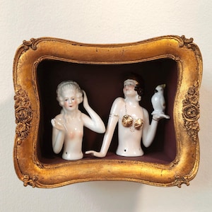Puede incluir: Una obra de arte enmarcada con dos bustos de muñecas de porcelana. Una muñeca tiene el pelo blanco, la otra tiene el pelo oscuro y un sujetador dorado. La segunda muñeca sostiene un pájaro blanco. El marco es dorado con detalles ornamentados.