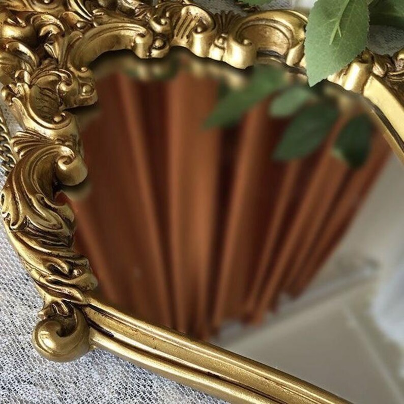 Close-up of an ornate gold Rococo mirror frame showing intricate Baroque scrollwork and filigree details, reflecting a warm terracotta orange curtain.