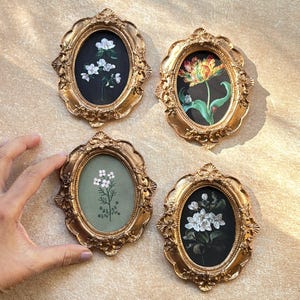May include: Four small, ornate, gold-coloured oval frames, each with a floral design. The flowers are in various colours against black and green backgrounds. The frames are arranged on a light-coloured surface.