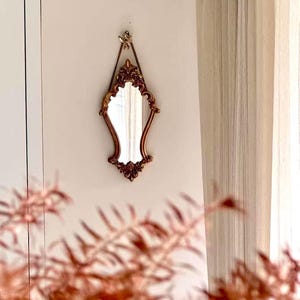 Vintage-style gold mirror with ornate baroque detailing, hanging by a chain from a decorative hook on a light-colored wall. Soft cream curtains are visible on the right, and delicate pinkish-brown fern leaves are blurred in the foreground.