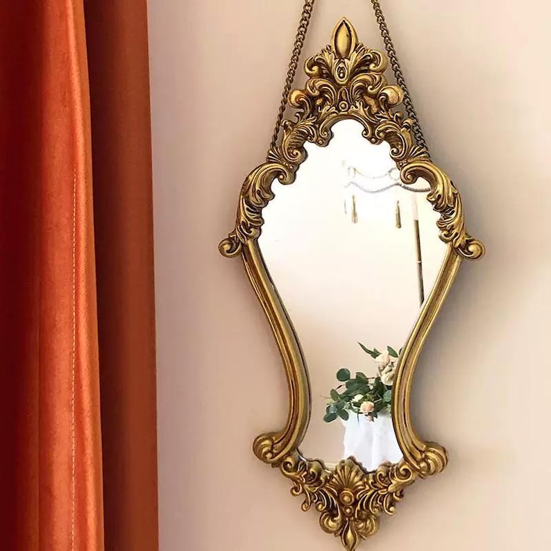 Ornate gold-framed vintage-style mirror hanging on a wall with a decorative chain. The mirror has an intricate baroque design and reflects a glimpse of green foliage and white flowers in the background. An orange curtain is partially visible to the left.