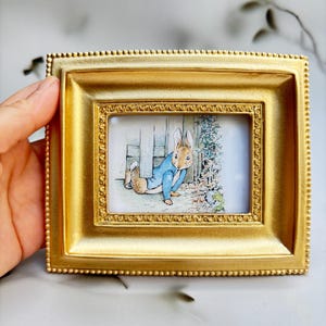 Op de afbeelding: Een kleine, rechthoekige goudkleurige fotolijst met een kralenrand. De ingelijste afbeelding toont een aquarelillustratie van een konijn in een blauw jasje. Het konijn staat in de buurt van een witte hek en groen.