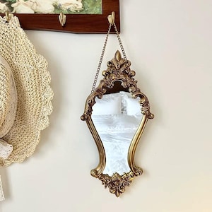 Vintage-style ornate gold mirror with an hourglass shape and floral carvings, hanging from a copper chain on a wall beside a straw hat.