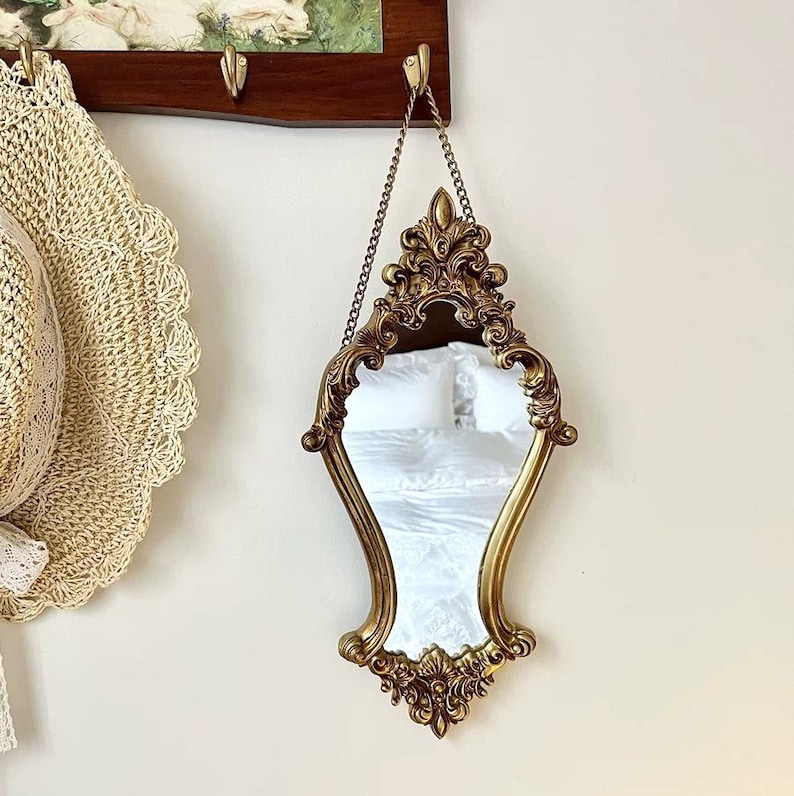Vintage-style ornate gold mirror with an hourglass shape and floral carvings, hanging from a copper chain on a wall beside a straw hat.