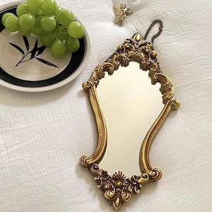 Decorative gold baroque mirror with carved floral details, displayed flat on white fabric beside a plate of green grapes and a small cherub charm.