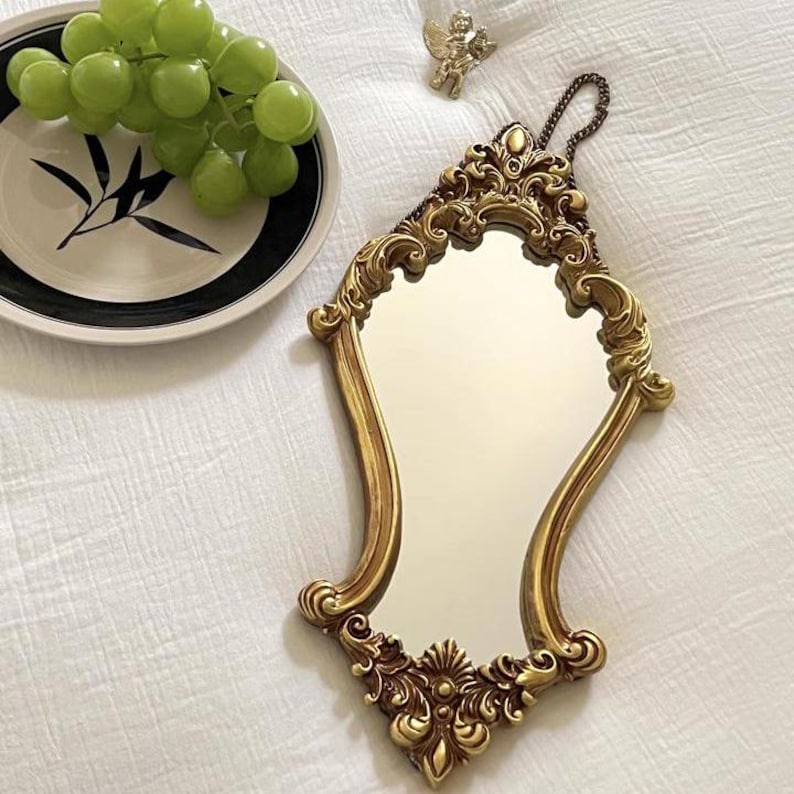 Decorative gold baroque mirror with carved floral details, displayed flat on white fabric beside a plate of green grapes and a small cherub charm.