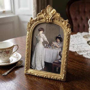 Könnte beinhalten: Vergoldeter, verzierter Bilderrahmen mit einem Vintage-Gemälde von zwei Frauen bei einer Teeparty. Der Rahmen hat ein detailliertes, geschwungenes Design. Eine Tasse und Untertasse mit floralen Mustern sind auf einem Holztisch zu sehen.