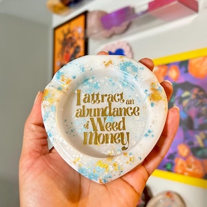 May include: White heart-shaped trinket dish with blue and gold glitter. The dish has the text "I attract an abundance of Weed money" in gold lettering.