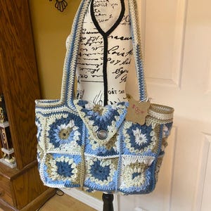 Könnte beinhalten: Eine gehäkelte Tragetasche in blau, weiß und braun. Die Tasche hat ein quadratisches Design mit einem Knopfverschluss. Das Preisschild zeigt "15,00 €".