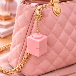 Puede incluir: Un bolso rosa acolchado con herrajes dorados, que incluye una correa de cadena y un dije en forma de corazón. Un pequeño accesorio rosa en forma de cubo cuelga de la cremallera. El bolso está sobre una tela rosa.