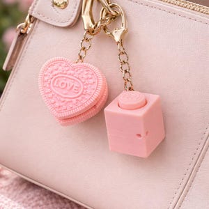 Puede incluir: Un bolso rosa claro con detalles dorados y dos colgantes rosas. Uno es una galleta en forma de corazón con la palabra "LOVE" grabada. El otro es un cubo rosa con un botón circular.