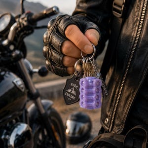 May include: A set of keys with a purple, cylindrical key holder and a black tag that reads "Ride Explore." The keys are held by a person wearing a black leather glove, with a motorcycle in the background.