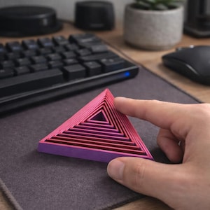 Puede incluir: Una pirámide geométrica en capas, en tonos rosa y morado, descansa sobre una superficie gris. La pirámide está compuesta por capas triangulares anidadas, creando un efecto 3D. Un dedo la toca.