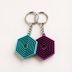 Puede incluir: Dos llaveros con colgantes hexagonales geométricos. Uno es turquesa, el otro morado. Ambos tienen un diseño en capas y están unidos a llaveros plateados con eslabones de cadena. Los llaveros se muestran sobre un fondo blanco.