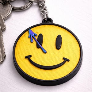 Puede incluir: Llavero de cara sonriente amarilla con borde negro y diseño de salpicaduras azules y moradas. El llavero está sujeto a una cadena plateada y un juego de llaves.